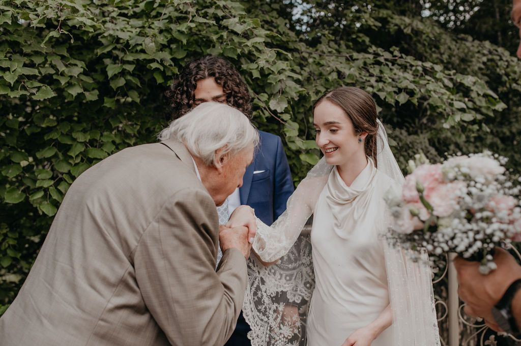 Elderly man kissing bride's hand at outdoor wedding ceremony while groom stands behind her.