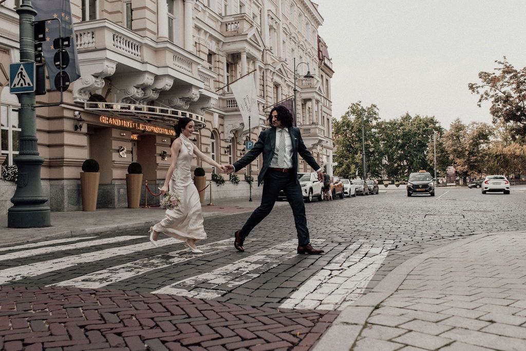 Bride and groom holding hands crossing a cobblestone street in an elegant European city during their wedding day.