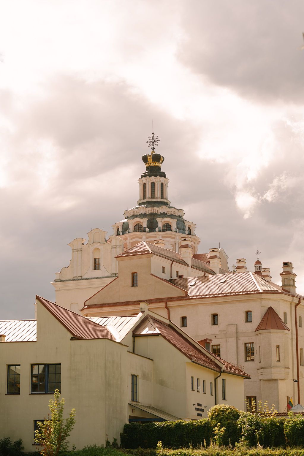 Historic baroque church with ornate dome and golden crown rises above white buildings under dramatic cloudy sky.