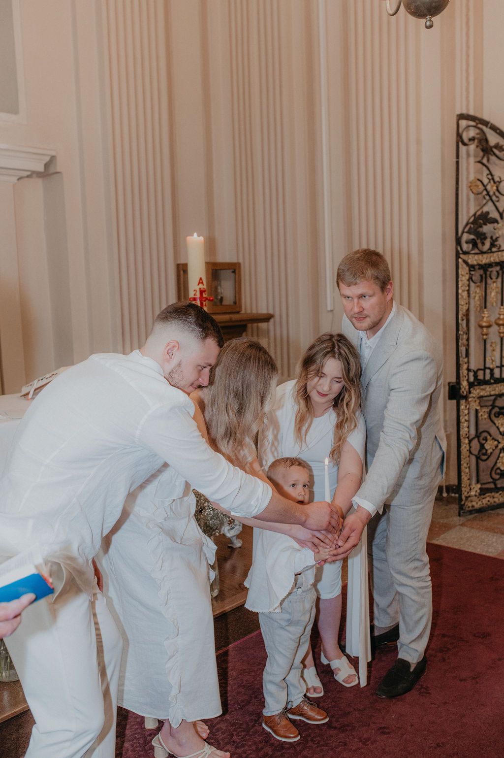 Family in white attire gathered around a baby during a christening ceremony in an elegant room with candles.