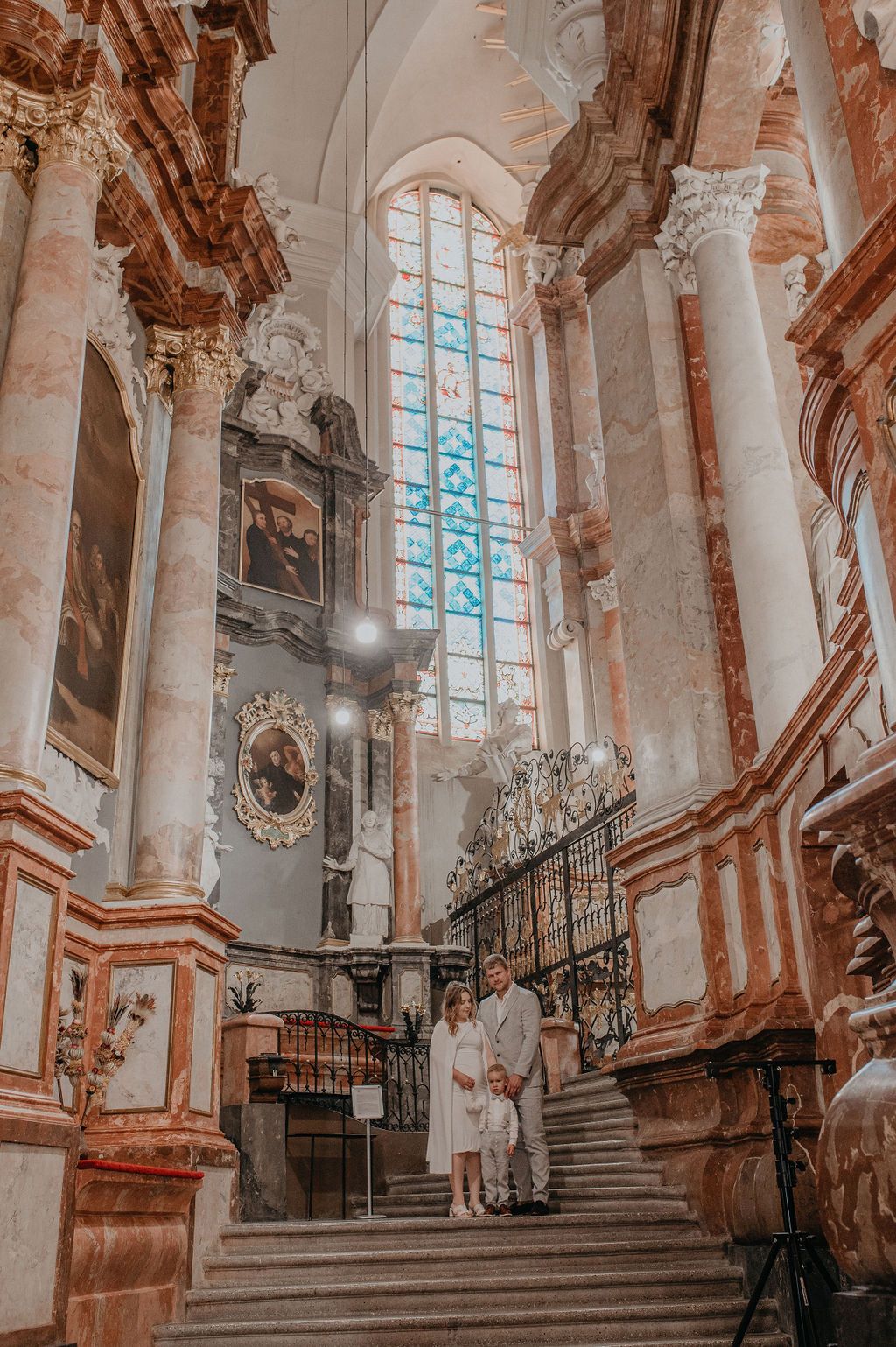 Family in white attire poses on ornate baroque church stairs with marble columns and stained glass windows.