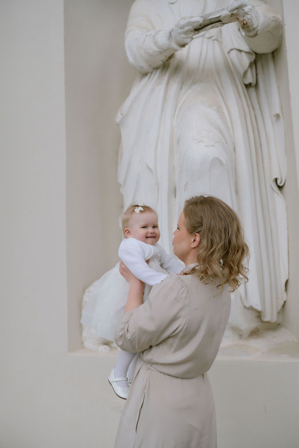 Mother holding smiling baby in white dress near classical marble statue in elegant interior.