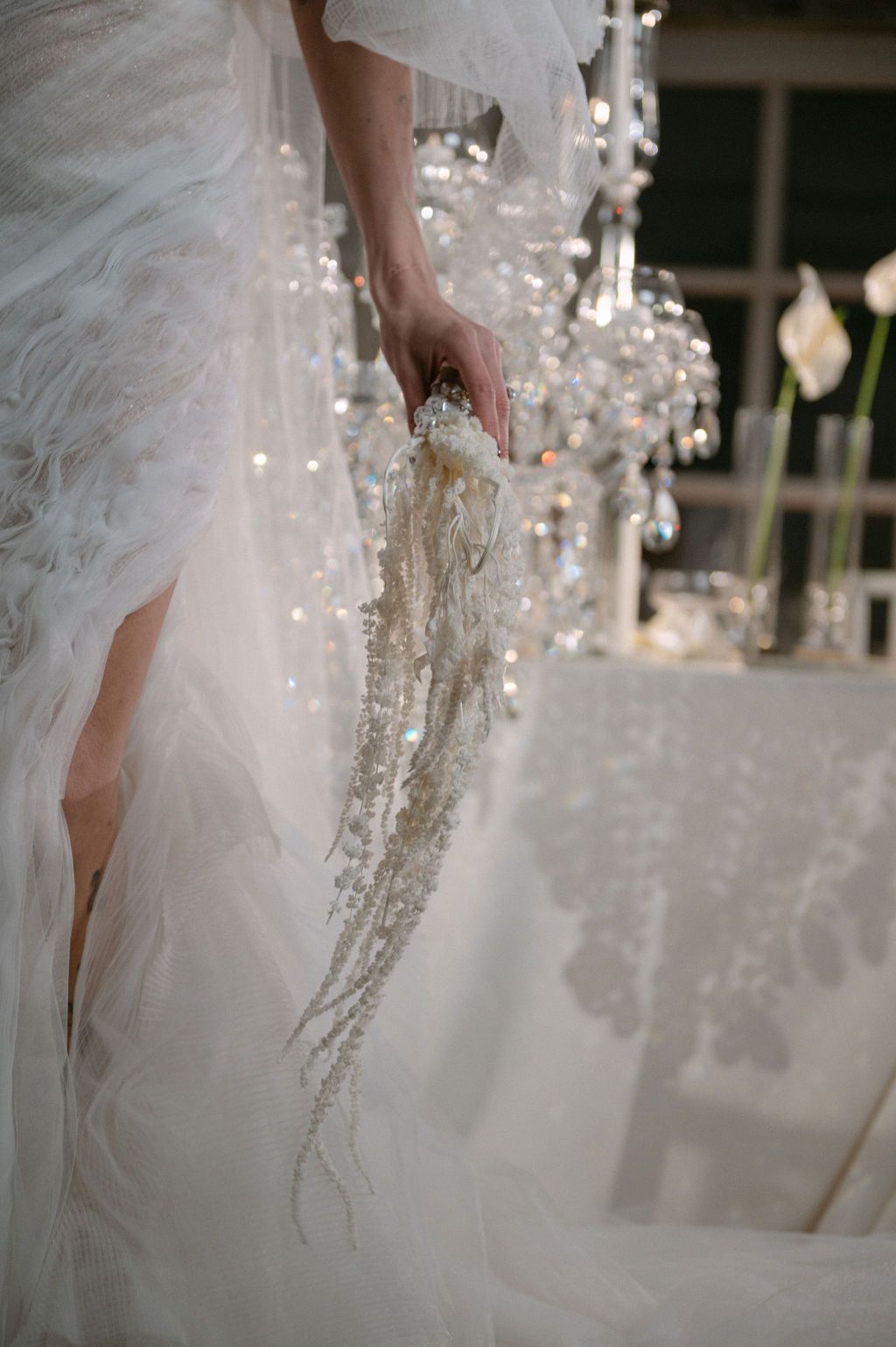 Bride in white feathered gown holding delicate pearl and crystal cascading bouquet near sparkling chandelier at elegant wedding.