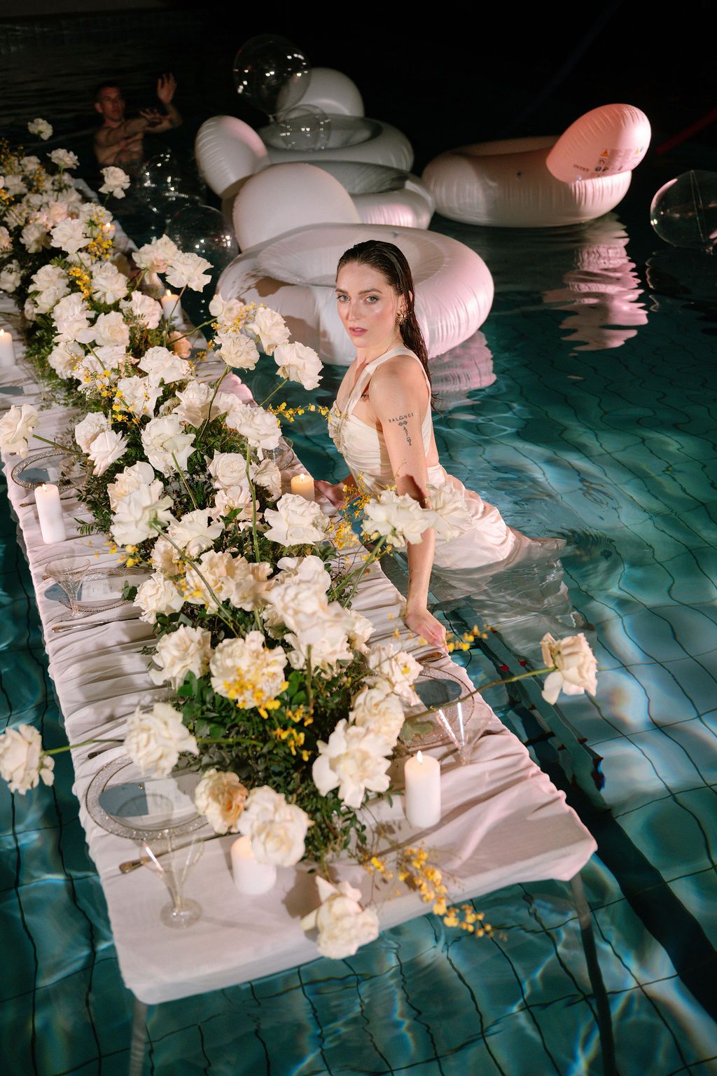 Woman in white dress standing in a pool beside a floating floral table with white flowers and candles.