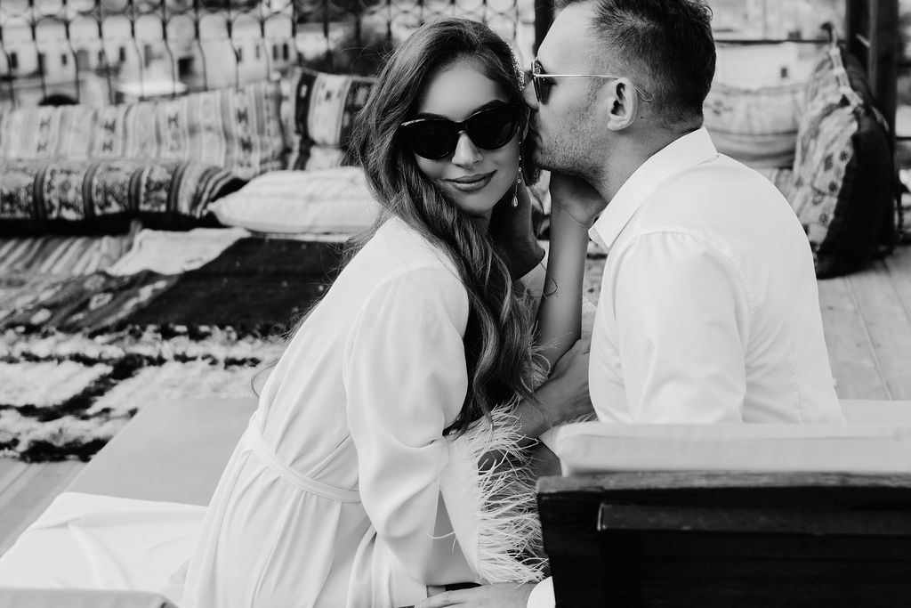 Black and white photo of a couple, man kissing woman's cheek, both in white outfits, woman wearing sunglasses.