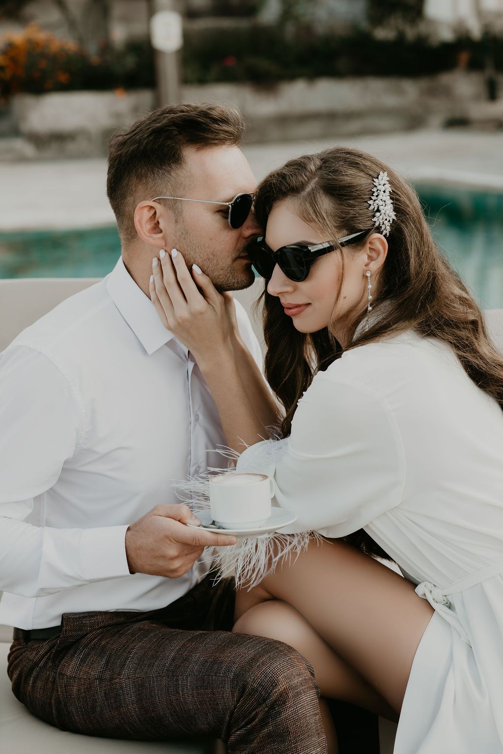 A romantic couple in sunglasses shares an intimate moment by a pool, woman in white dress holding a coffee cup.