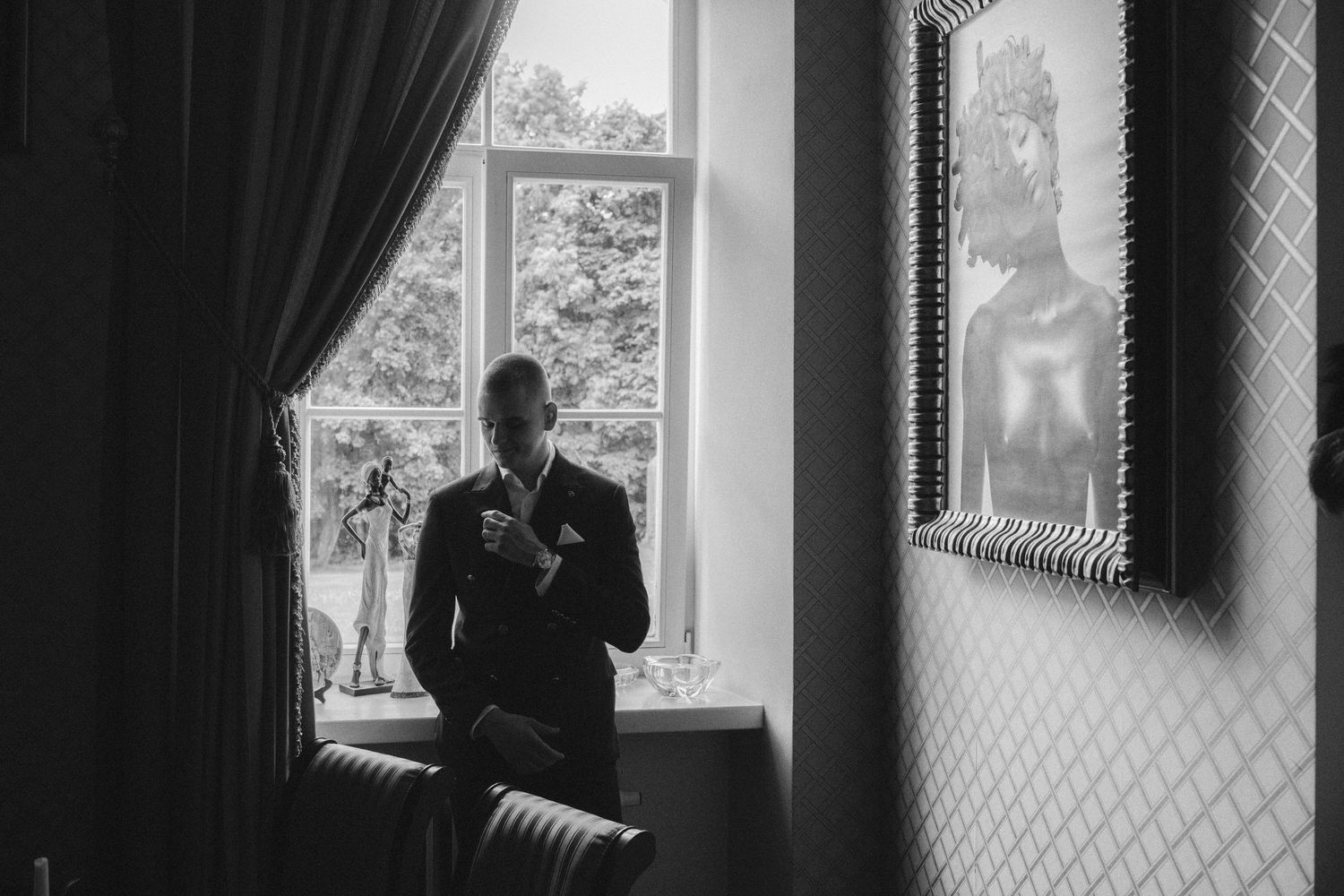 Black and white photo of a groom in a suit adjusting his tie by a window on his wedding day.