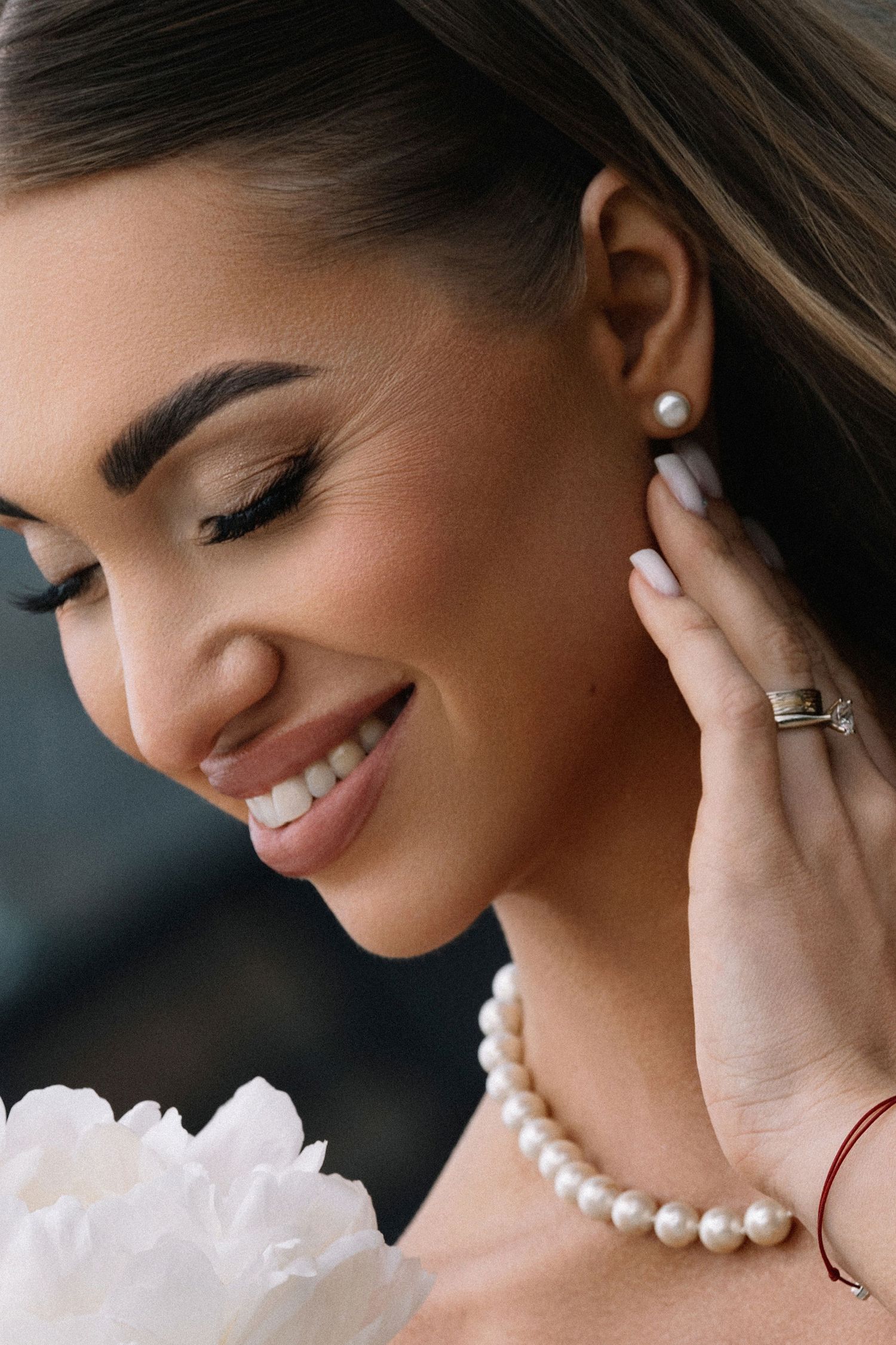 Smiling bride wearing pearl necklace, earrings, and ring, touching her ear while holding white peony flowers.