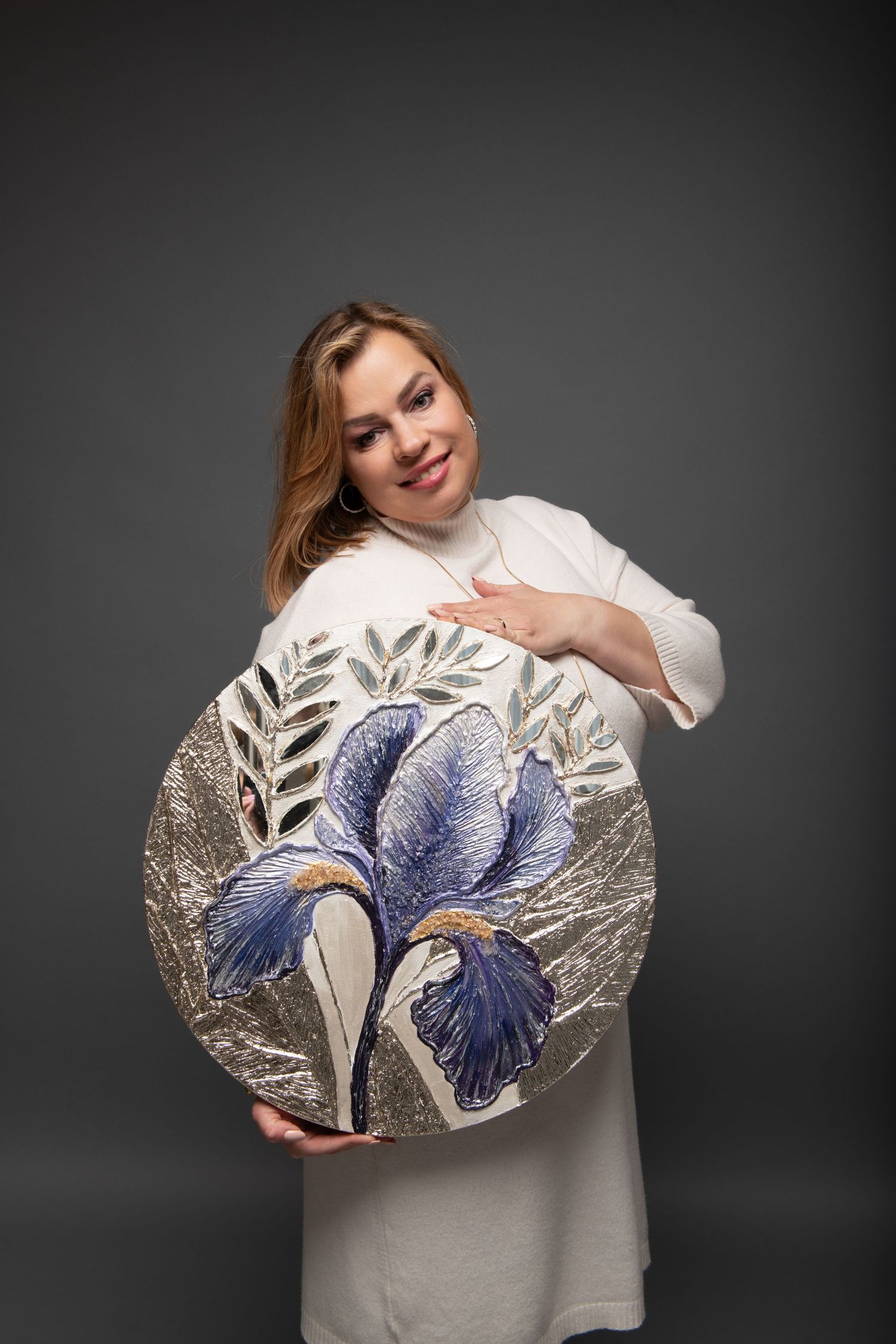 Woman in white dress holding a decorative round ceramic plate with blue iris flower and silver leaf design.