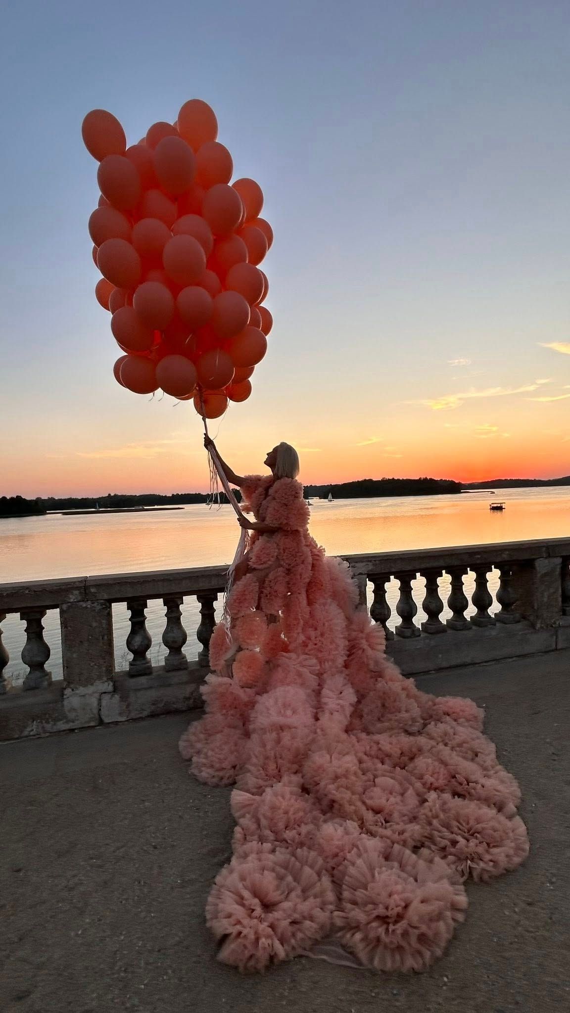Woman in pink ruffled gown holding red balloons at sunset by a waterfront balustrade.