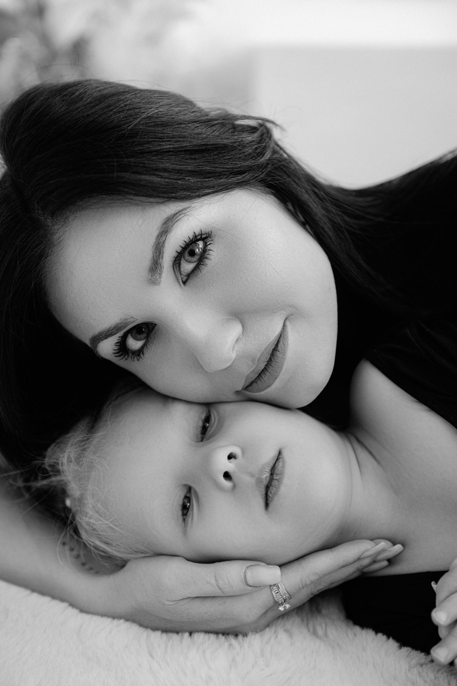 Black and white portrait of a mother tenderly resting her cheek on her young child's head, both gazing at the camera.