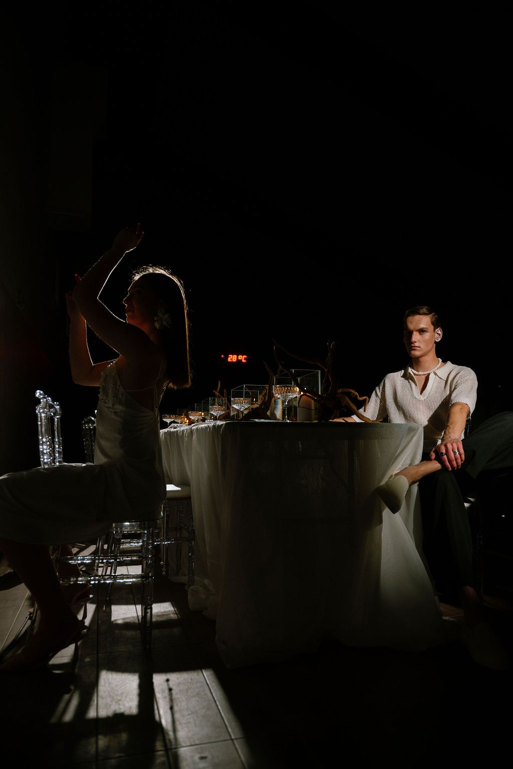 A man in a white shirt sits at a draped table in a dark room while a woman stands in dramatic light, raising her arm.