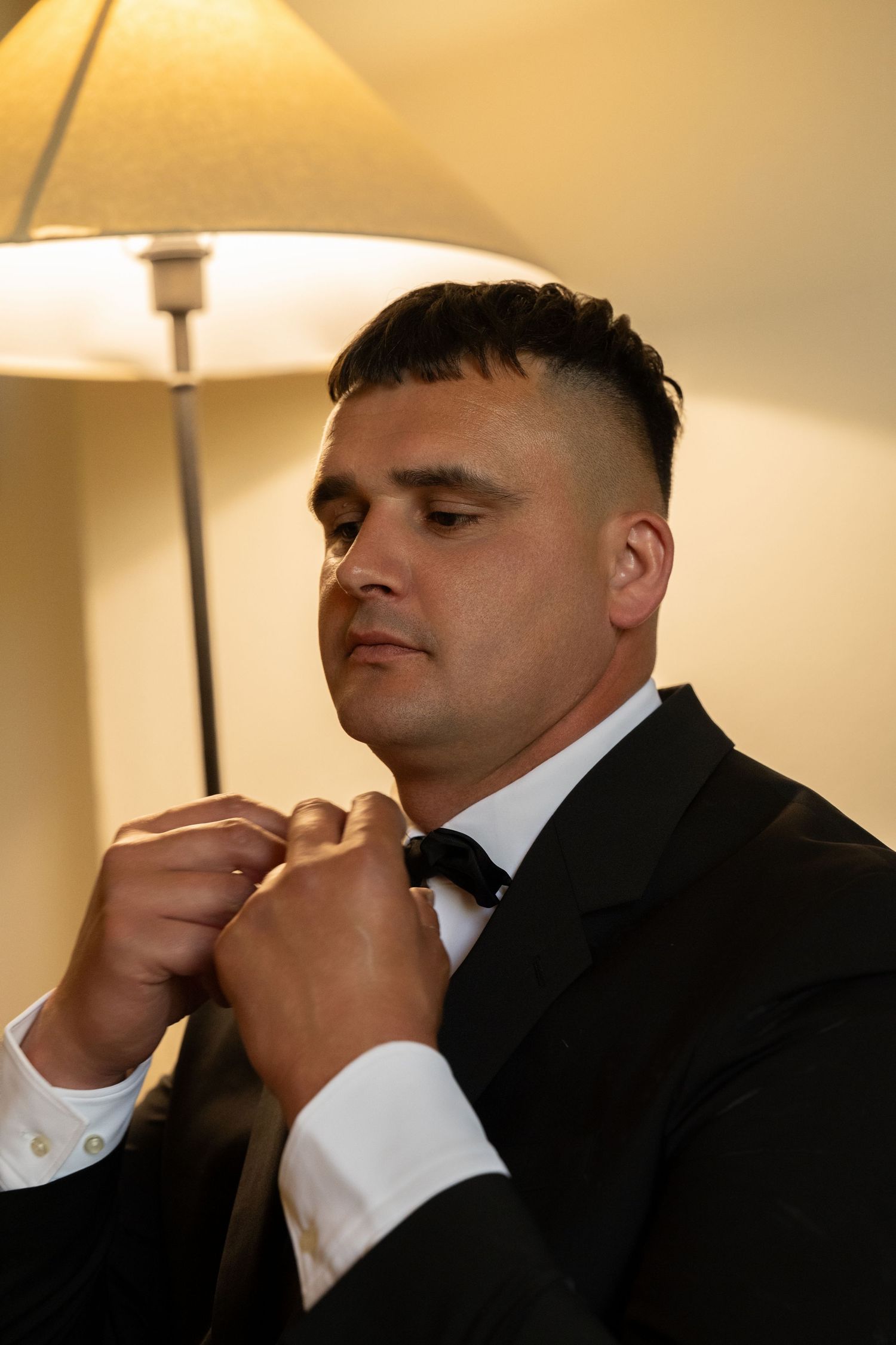 Groom with a short fade haircut adjusts his black bow tie in a dark suit beside a glowing lamp before the wedding.