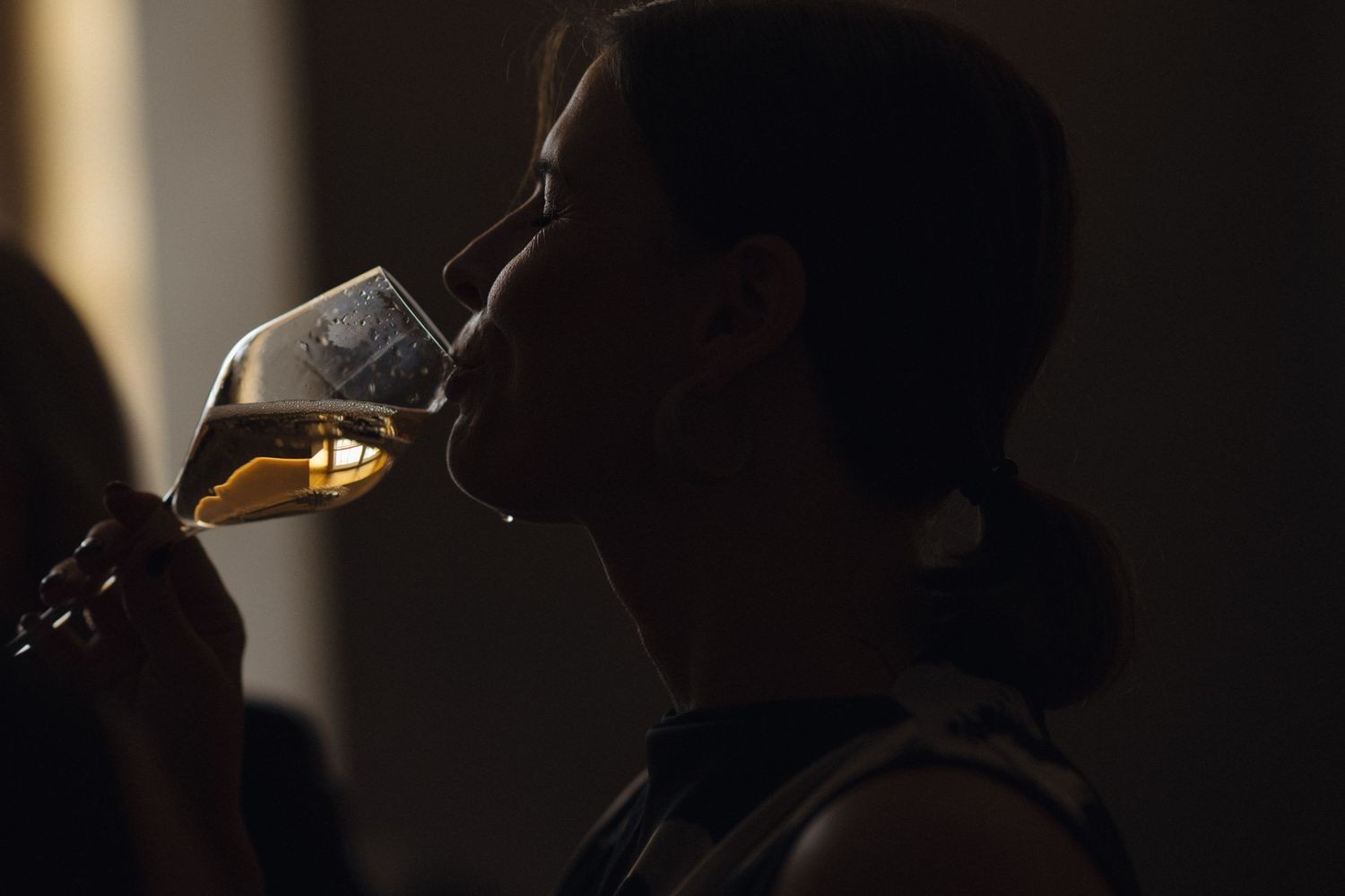 Woman in silhouette with hair pulled back sipping golden white wine from a stemmed glass in a dimly lit room.