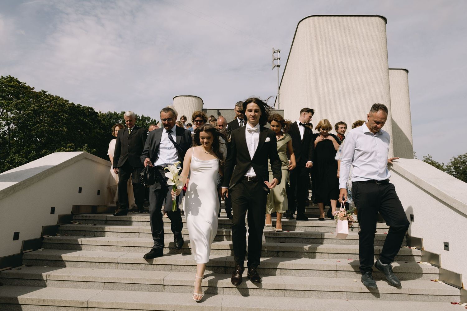 Bride in a white dress and groom in a dark suit walk down outdoor concrete steps with smiling wedding guests following behind them.