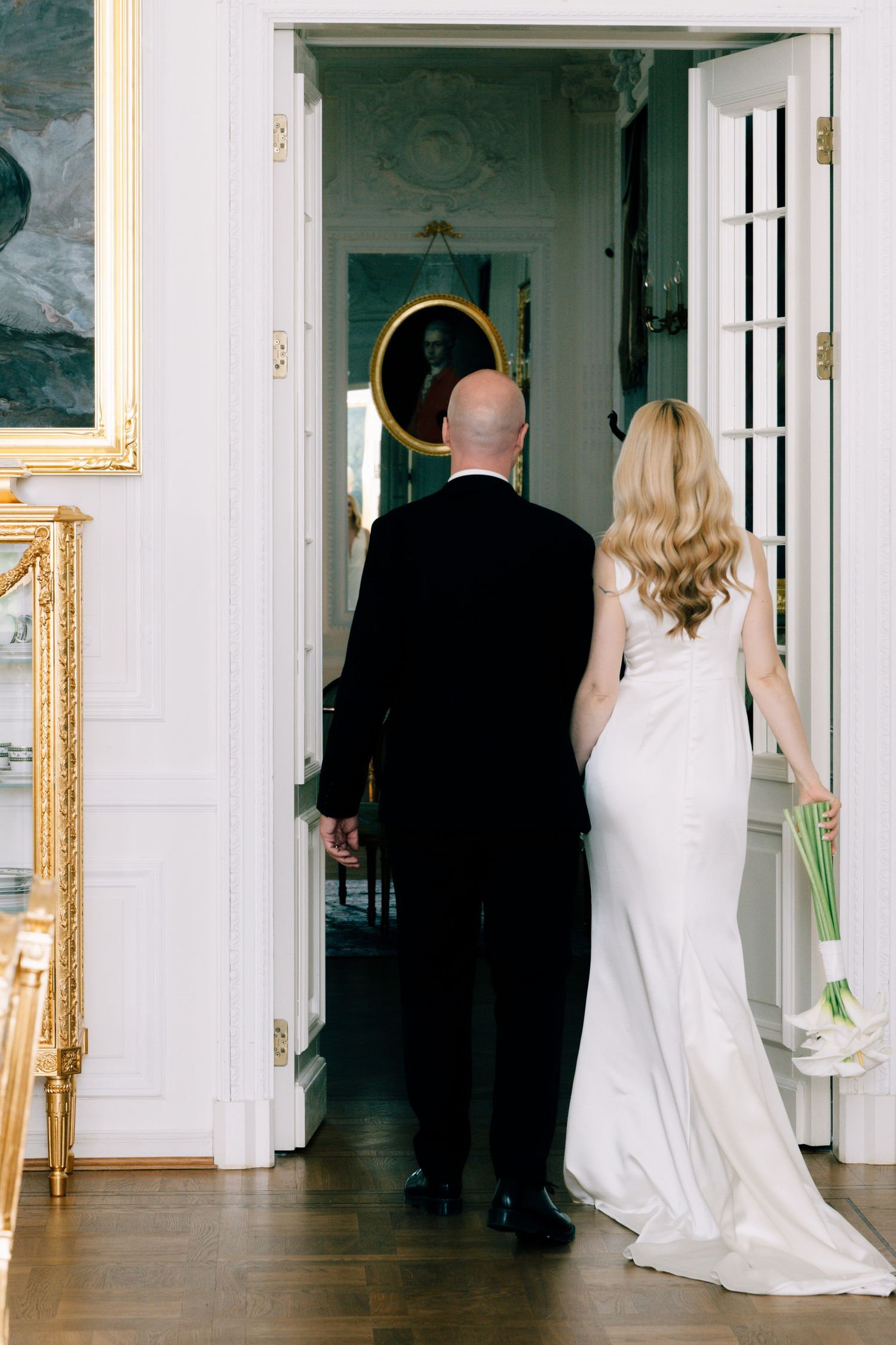 A bride in a white gown holding a white calla lily bouquet walks with her groom in a black suit through an elegant white doorway.