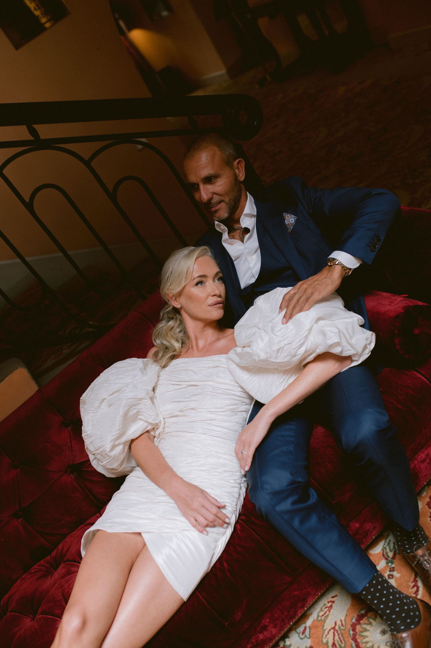 A couple poses intimately on a red velvet couch in a romantic boudoir photography session.
