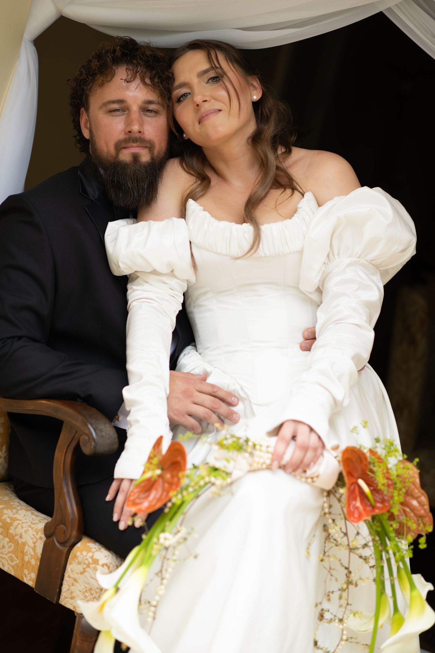 Bride in white off-shoulder gown with puff sleeves holds floral bouquet beside bearded groom in black suit.