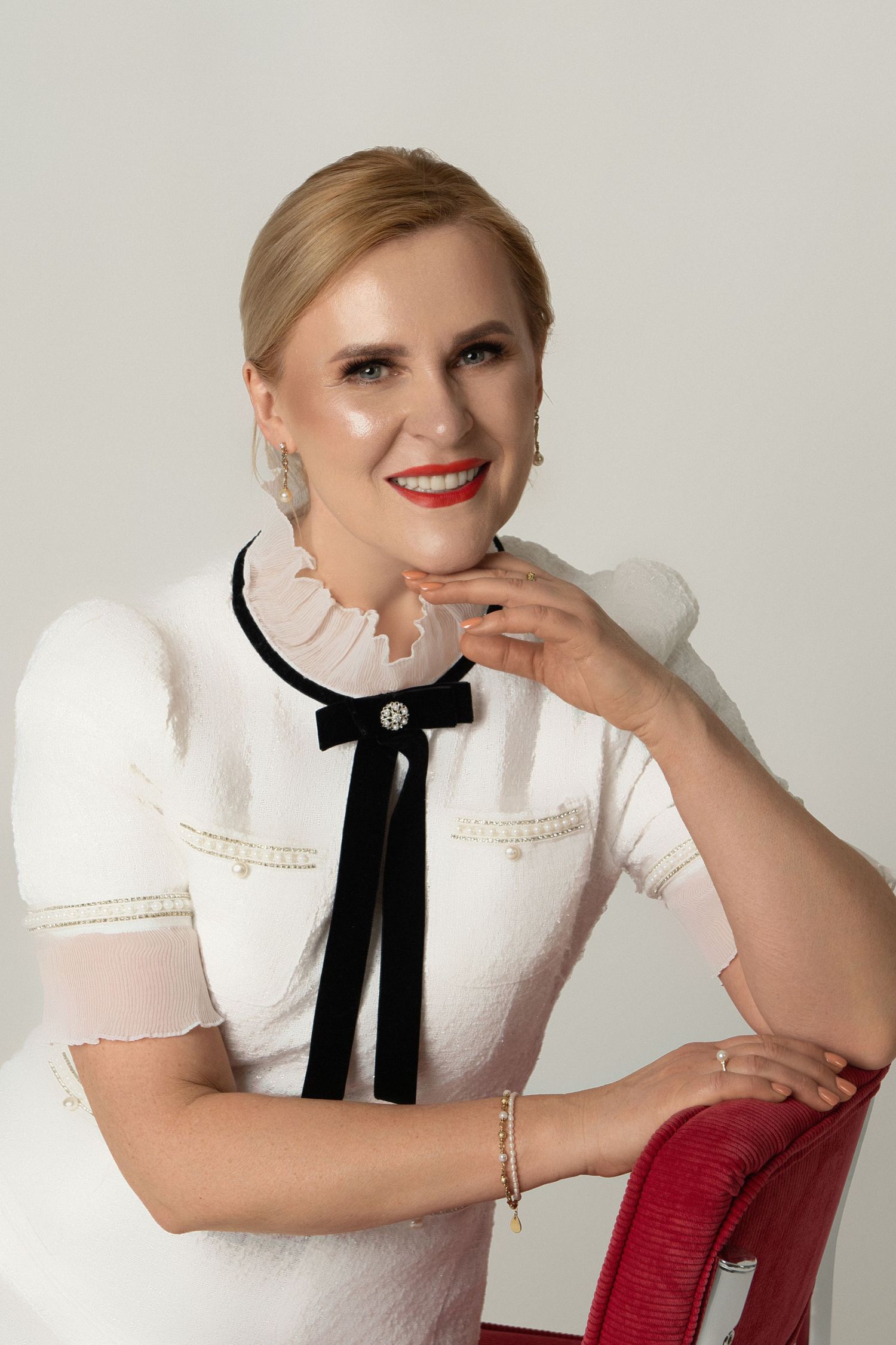 Smiling blonde woman in white dress with black bow, red lips, pearl earrings, seated elegantly in studio portrait.