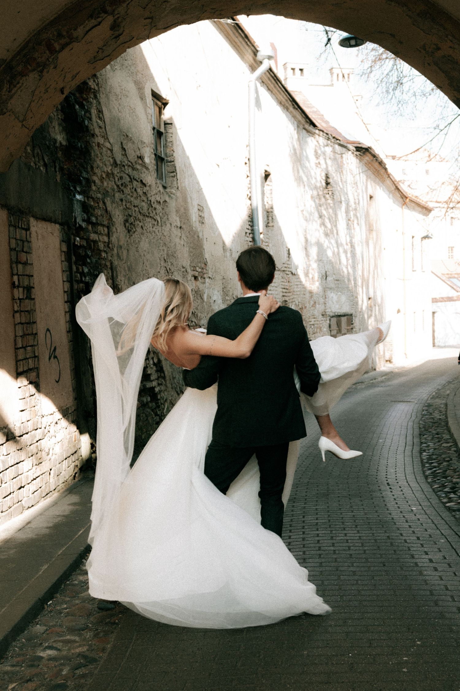 A groom in a black suit lifts his bride in a white gown through a historic cobblestone archway.