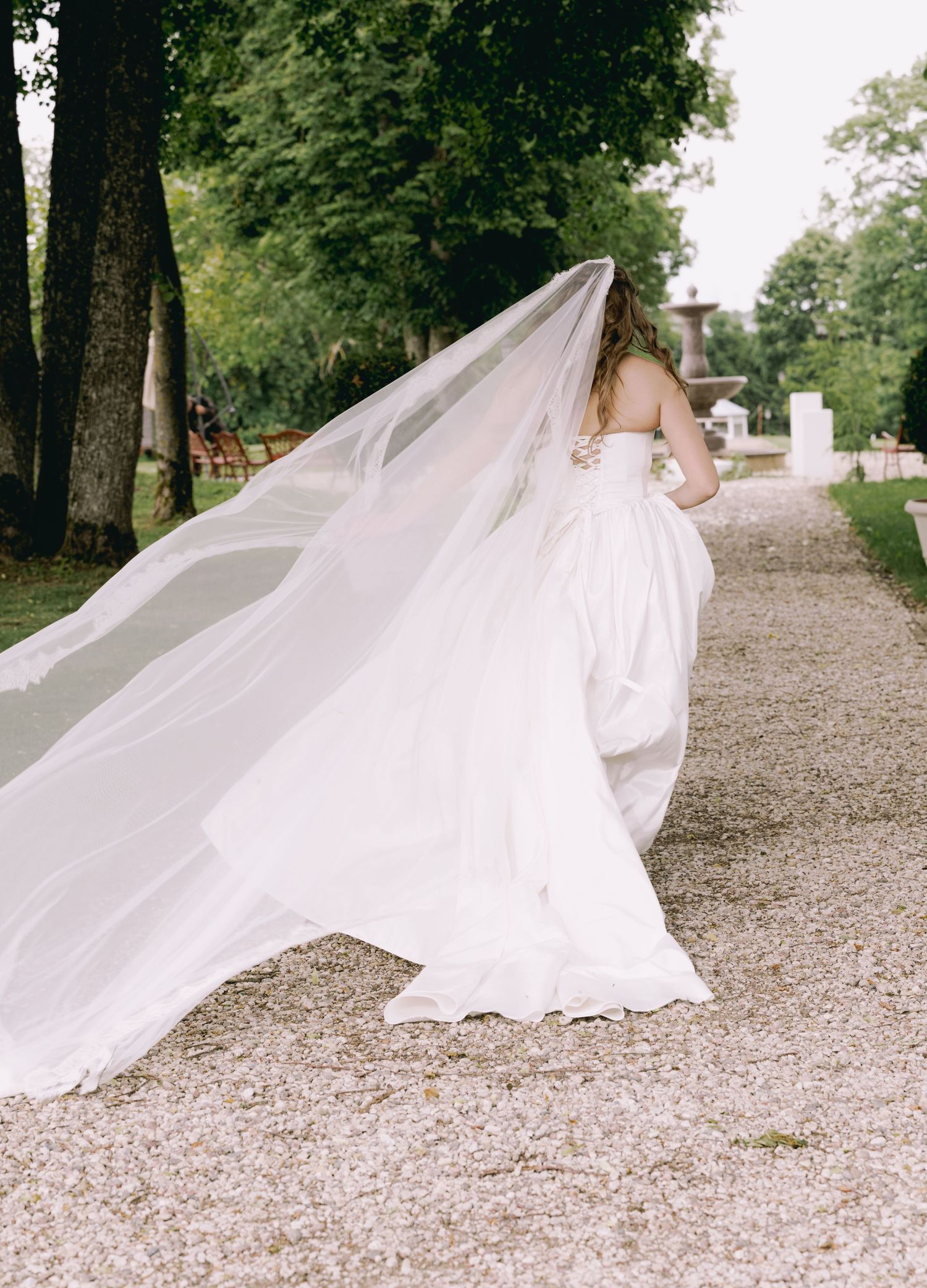 Bride in flowing white gown and long veil walks gracefully along tree-lined gravel path.