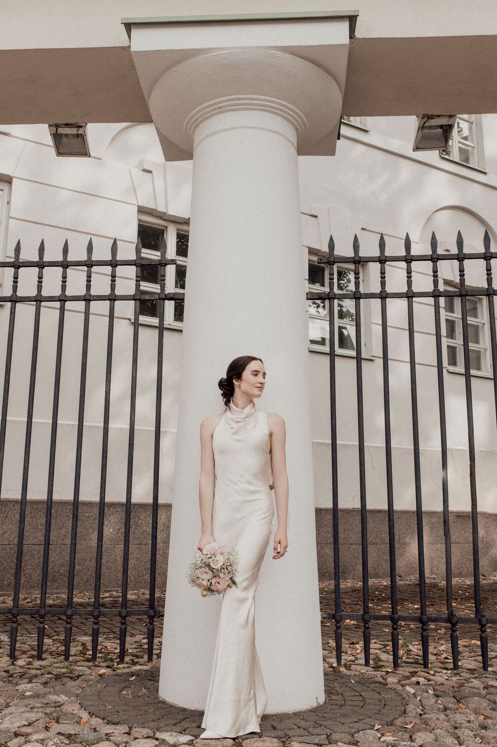 Bride in elegant white satin gown holding bouquet, posing beside a white column with iron fence backdrop.