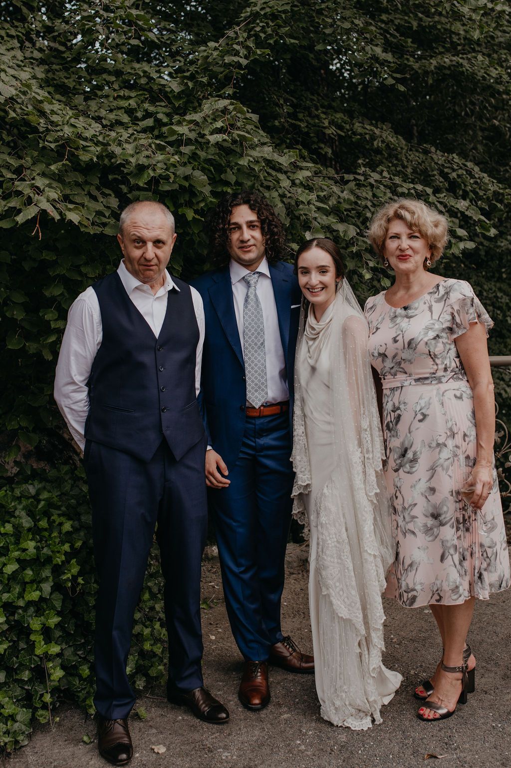 Wedding family portrait of four people posing outdoors in front of green foliage.