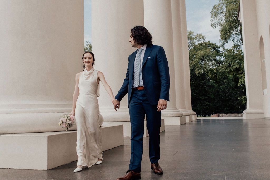 Bride and groom hold hands walking between white columns in an elegant outdoor wedding portrait.
