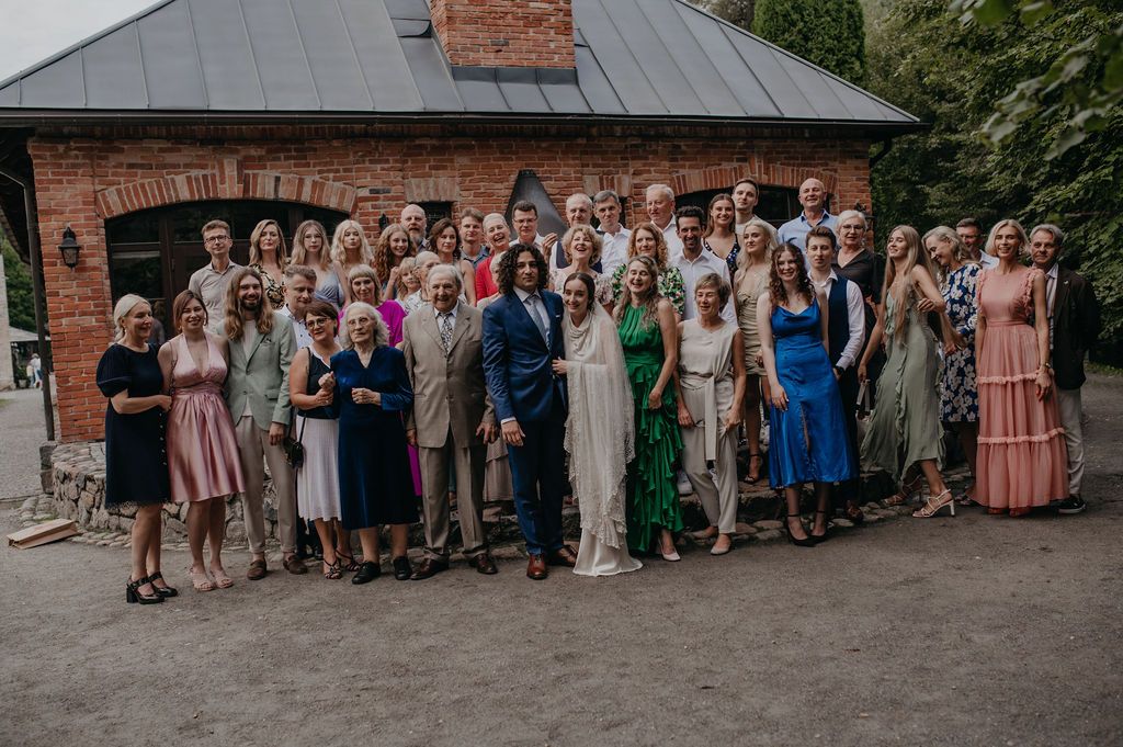 Large wedding party group photo outside rustic brick building, guests in formal attire surrounding bride and groom.