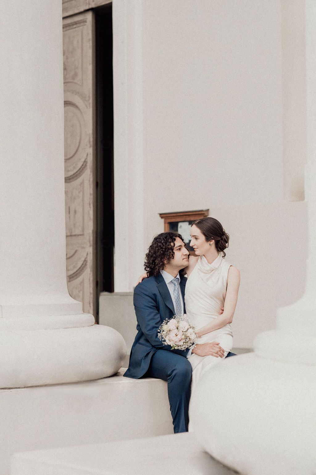 Bride and groom sit closely together on white stone steps, sharing an intimate moment with a bridal bouquet.