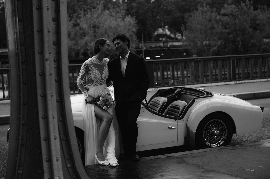 Black and white photo of a bride and groom posing beside a classic white sports car outdoors.