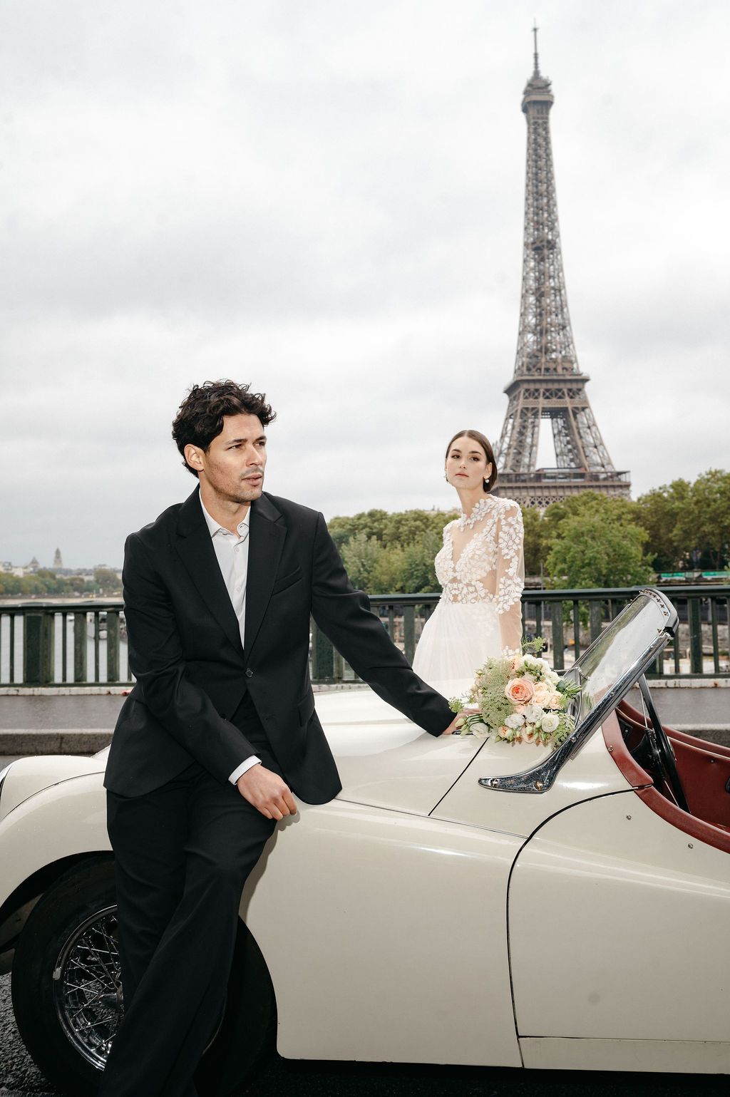 A bride and groom pose with a vintage white convertible car in front of the Eiffel Tower in Paris.
