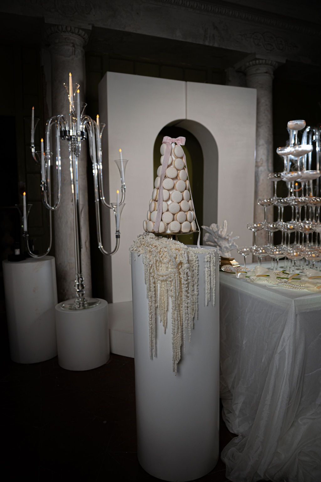 Elegant wedding dessert display with macaron tower, crystal candelabras, and champagne glass pyramid on white pedestals.
