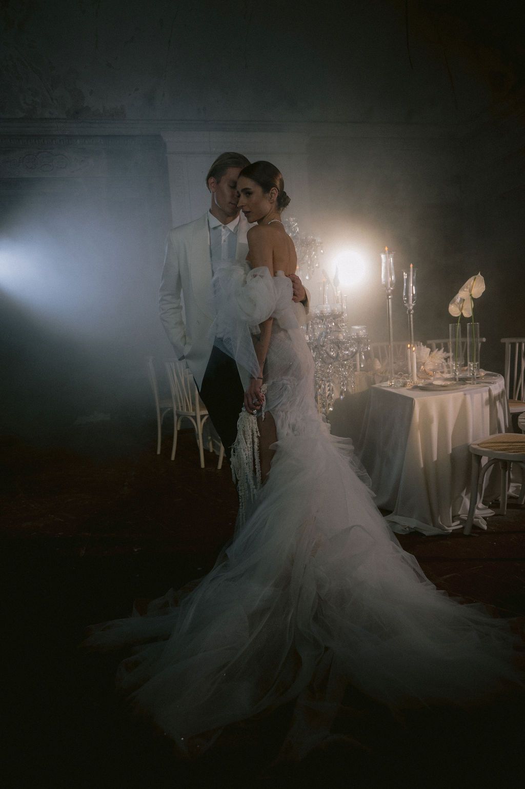 Bride in mermaid gown and groom in white tuxedo share romantic moment in dramatic candlelit wedding venue.