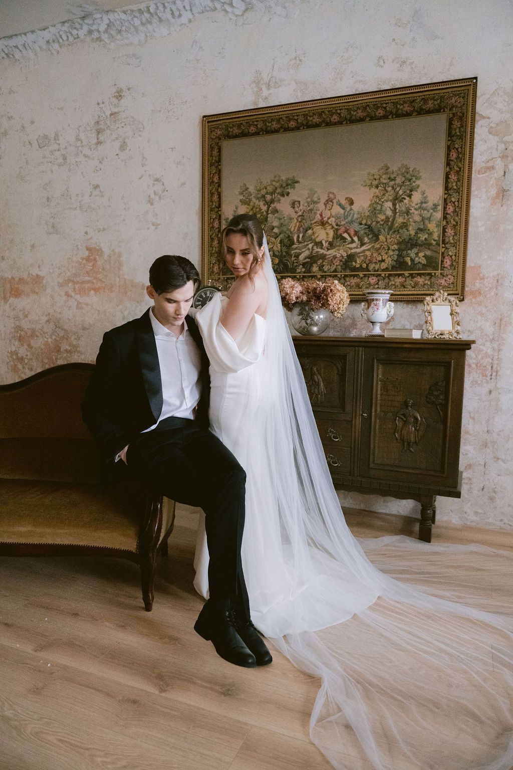 Bride in white gown with long veil stands beside seated groom in black tuxedo near ornate vintage cabinet.