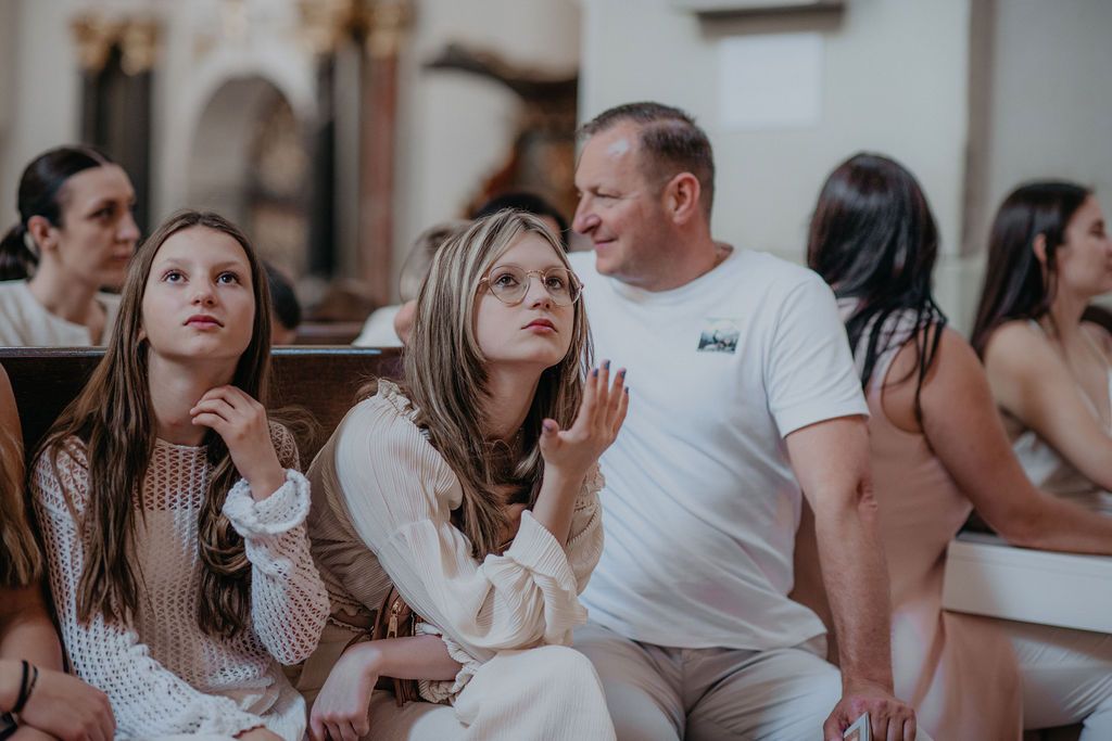 Family sitting in church pews, two girls and a man in white shirt looking upward during a religious ceremony.