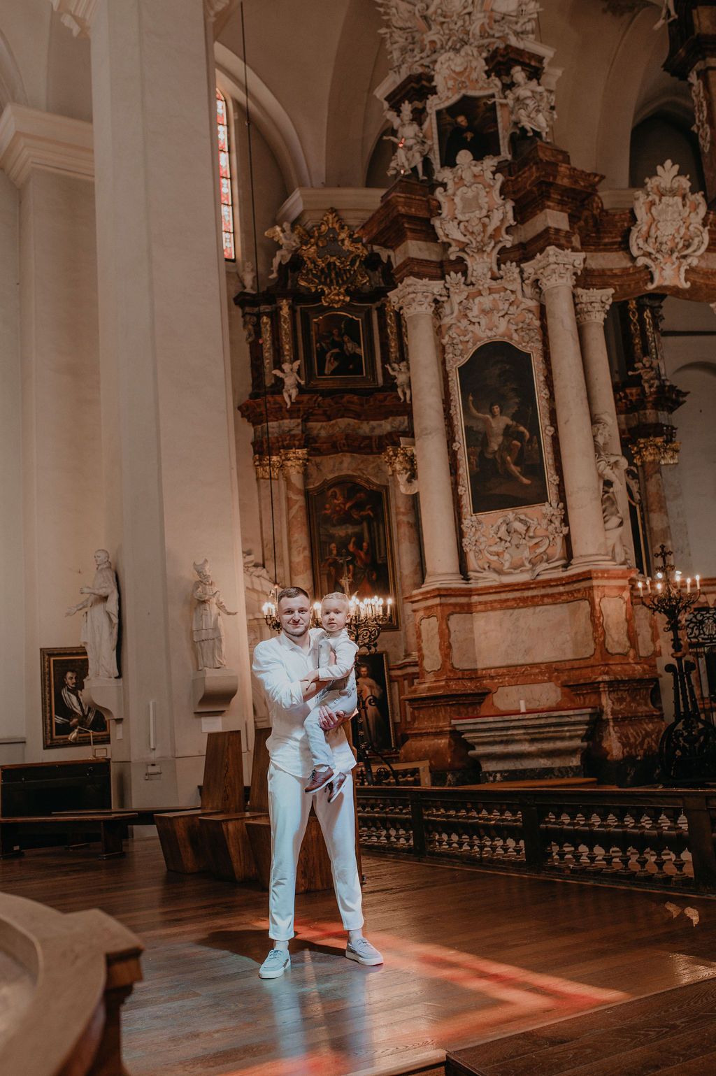 Man in white suit holding a baby in a ornate baroque church interior with golden altar.
