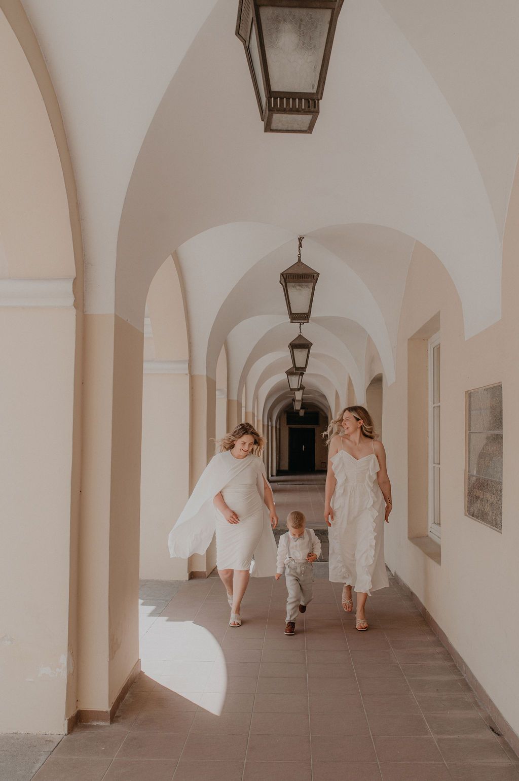 Two women in white dresses walk with a child through an elegant arched corridor with hanging lanterns.