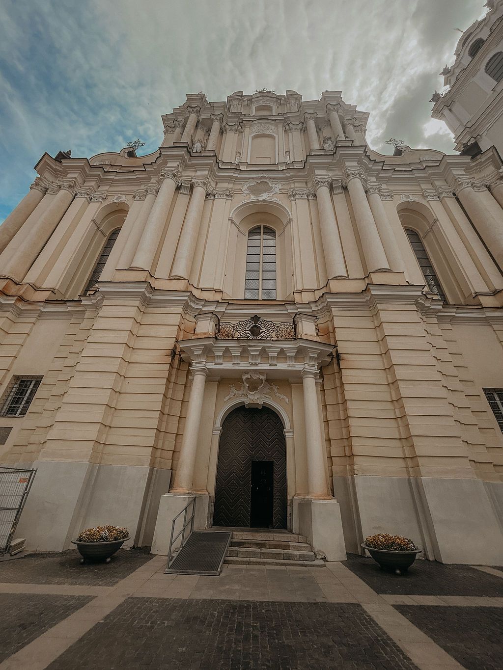 Baroque church facade with ornate columns, arched entrance, and decorative stonework under a cloudy sky.