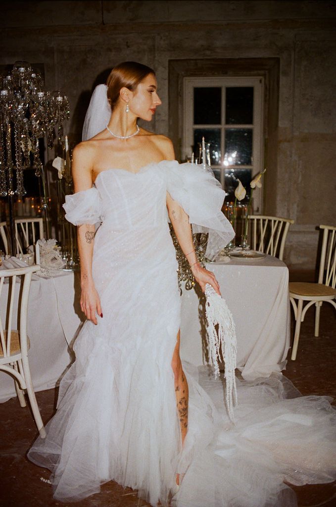 Bride in off-shoulder white tulle wedding gown with veil and tattoos, holding bouquet in elegant reception venue.