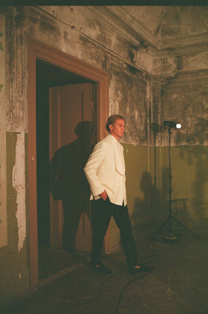 Man in white blazer standing in dimly lit, weathered room with studio light and shadow on wall.