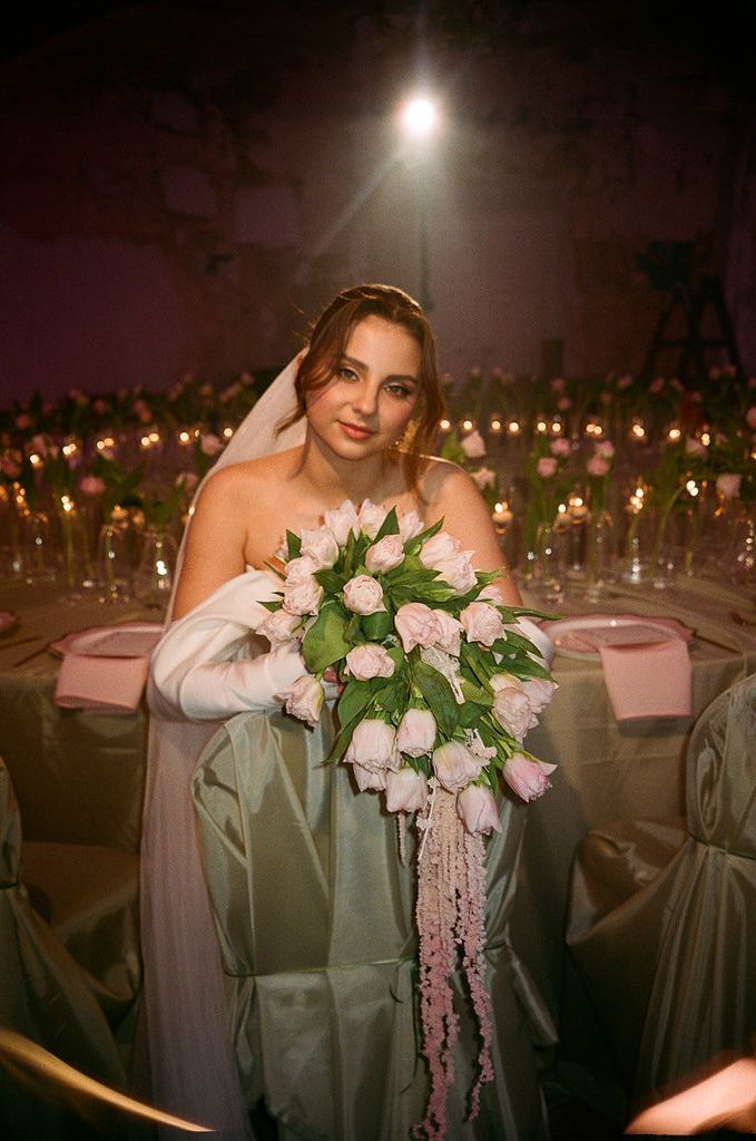 Bride in white veil holding cascading pink tulip bouquet seated at elegantly decorated wedding reception table.