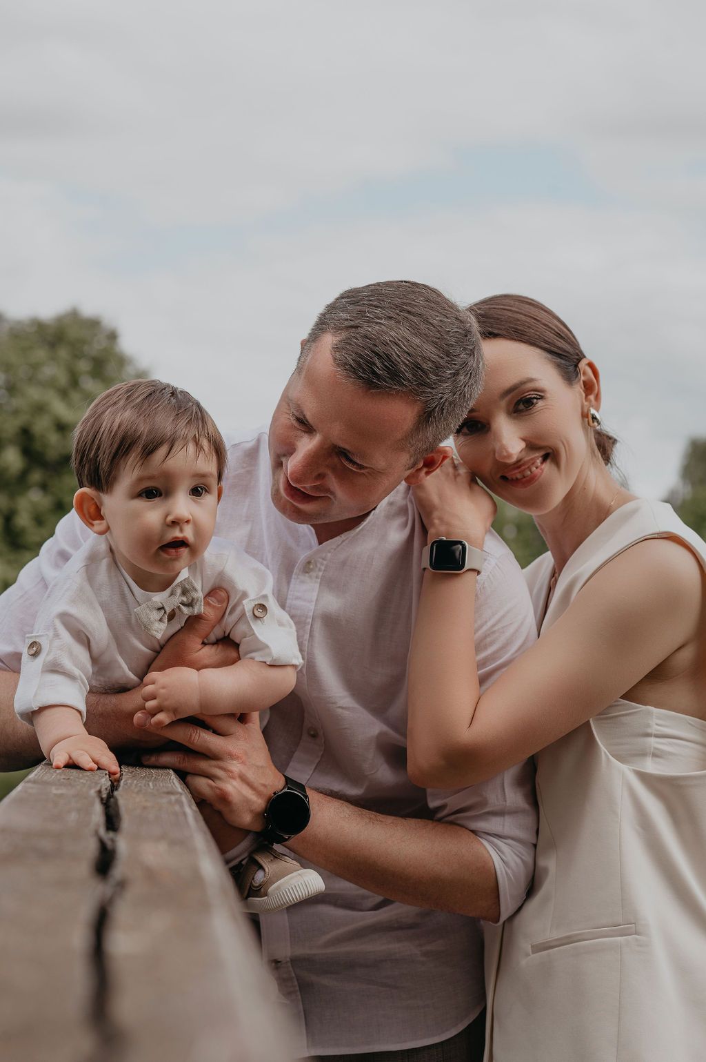 A happy young family with a baby boy in white outfits poses outdoors near a wooden railing on a cloudy day.