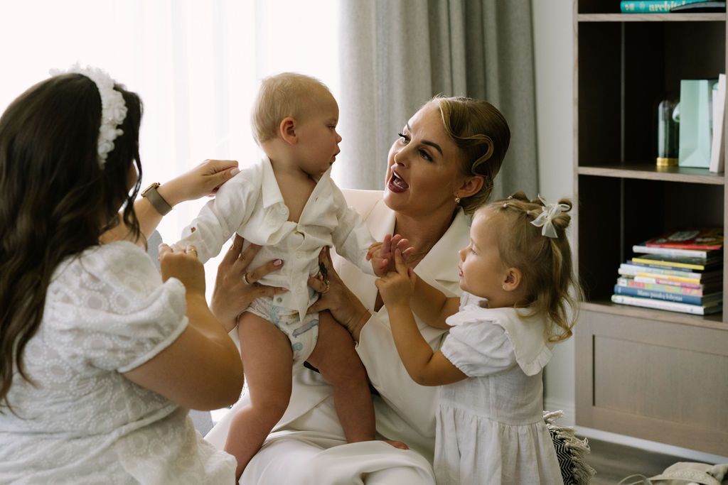 A woman in white holds a baby while two young girls in white dresses interact joyfully in a bright, cozy room.