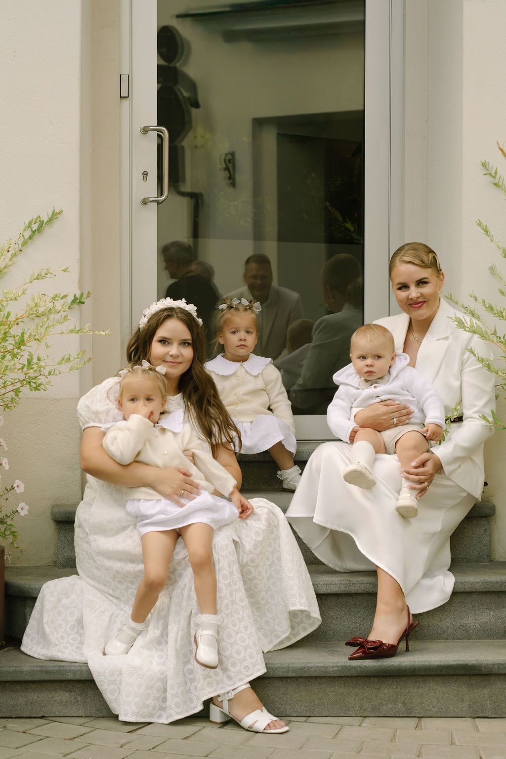 Two women in white dresses sit on steps with three young children during a formal christening ceremony.