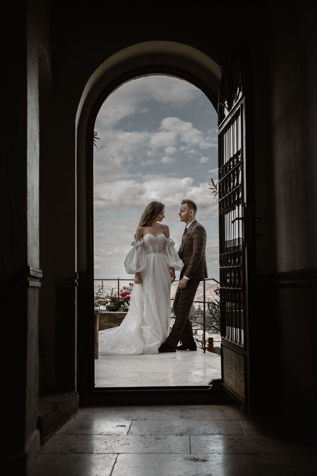Bride and groom standing in an arched doorway with a cloudy sky backdrop on their wedding day.