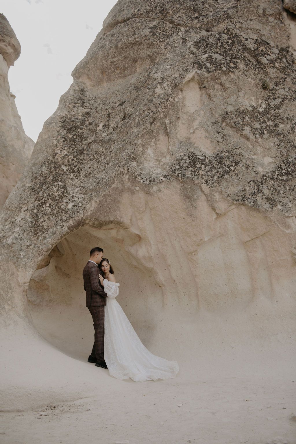 Bride and groom embracing beneath dramatic sandstone rock formations in Cappadocia, Turkey, wedding photo.