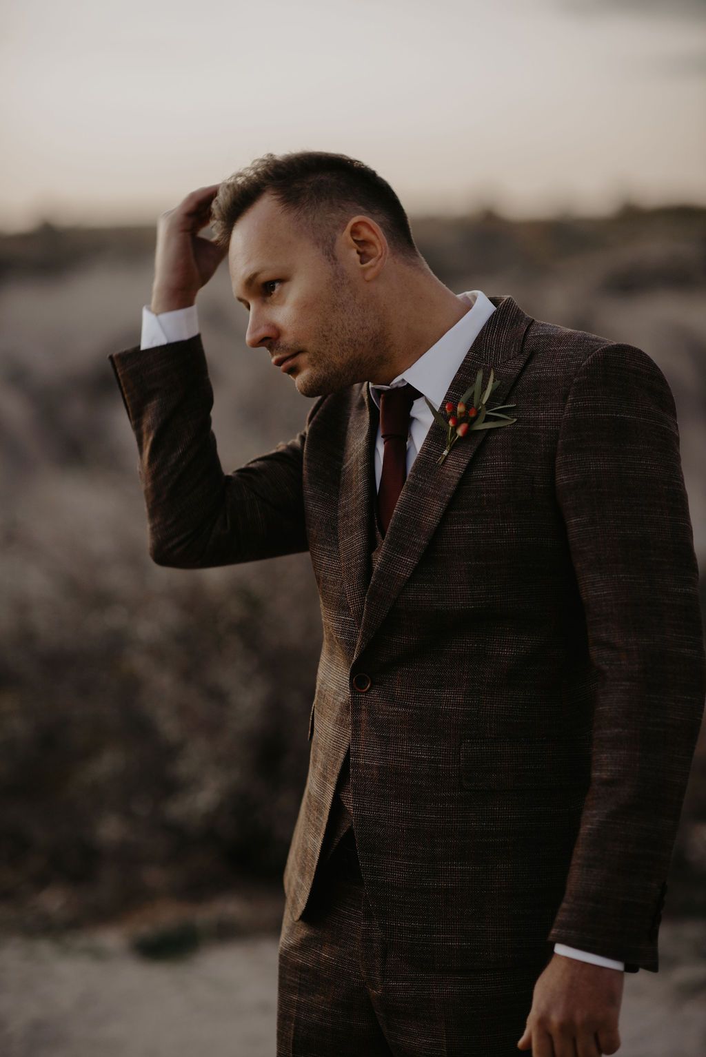 Groom in brown plaid suit with boutonniere running hand through hair at outdoor wedding.