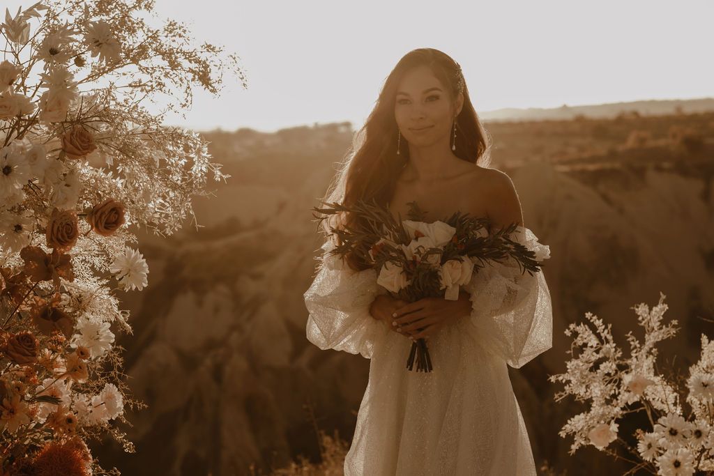 Bride in off-shoulder white gown holds bouquet amid warm golden sunset in rocky terrain.