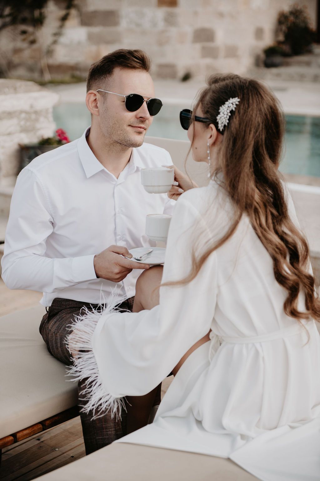 Elegant couple in white outfits sharing coffee by a pool, wearing stylish sunglasses in a romantic outdoor setting.